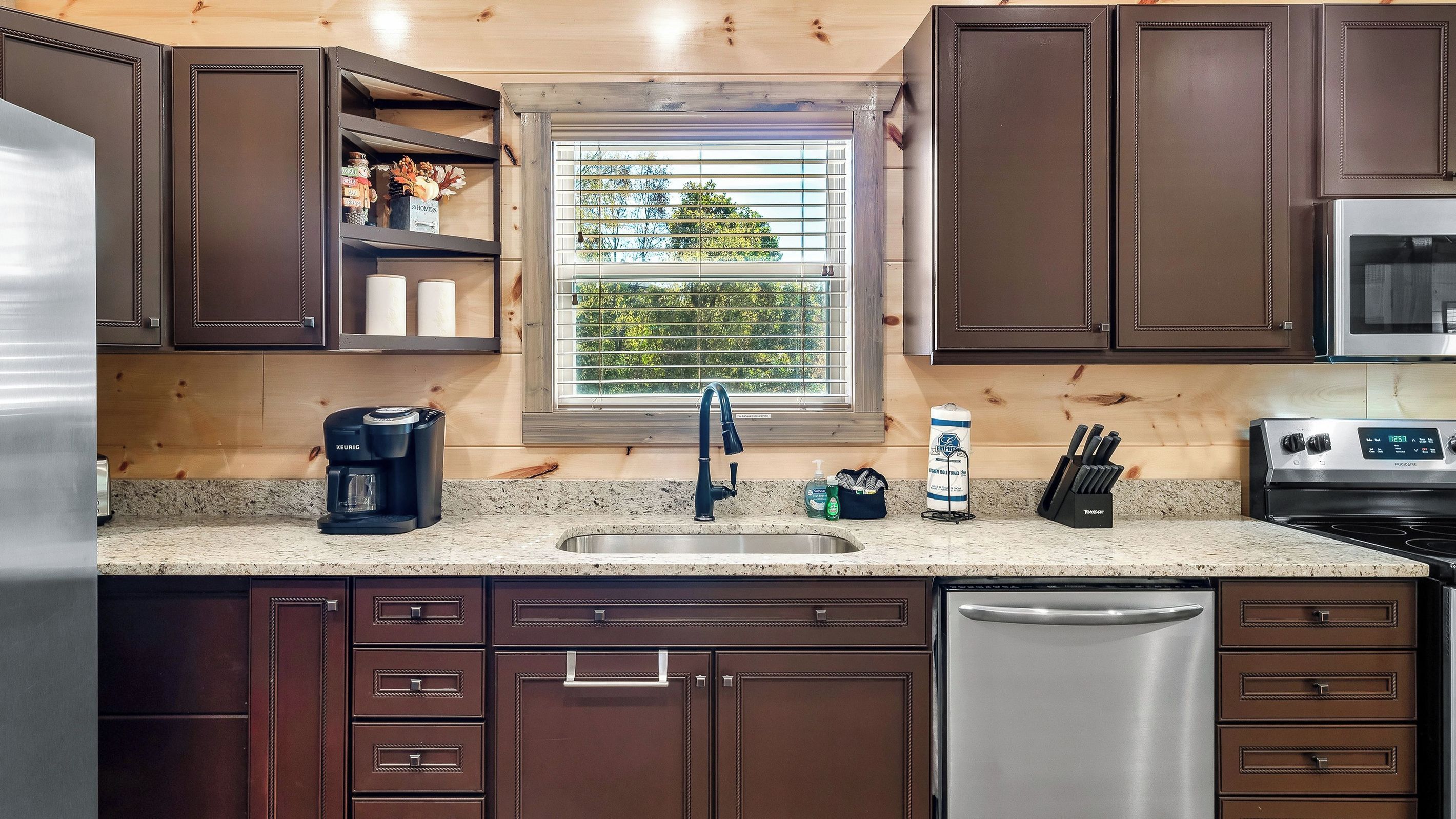 Una cocina con armarios marrón oscuro, un refrigerador de acero inoxidable, una cafetera, un lavamanos y un ventanal con vistas a árboles.