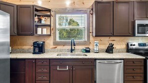 A kitchen with dark brown cabinets, a stainless steel refrigerator, a coffee maker, a sink, and a window with a view of trees.