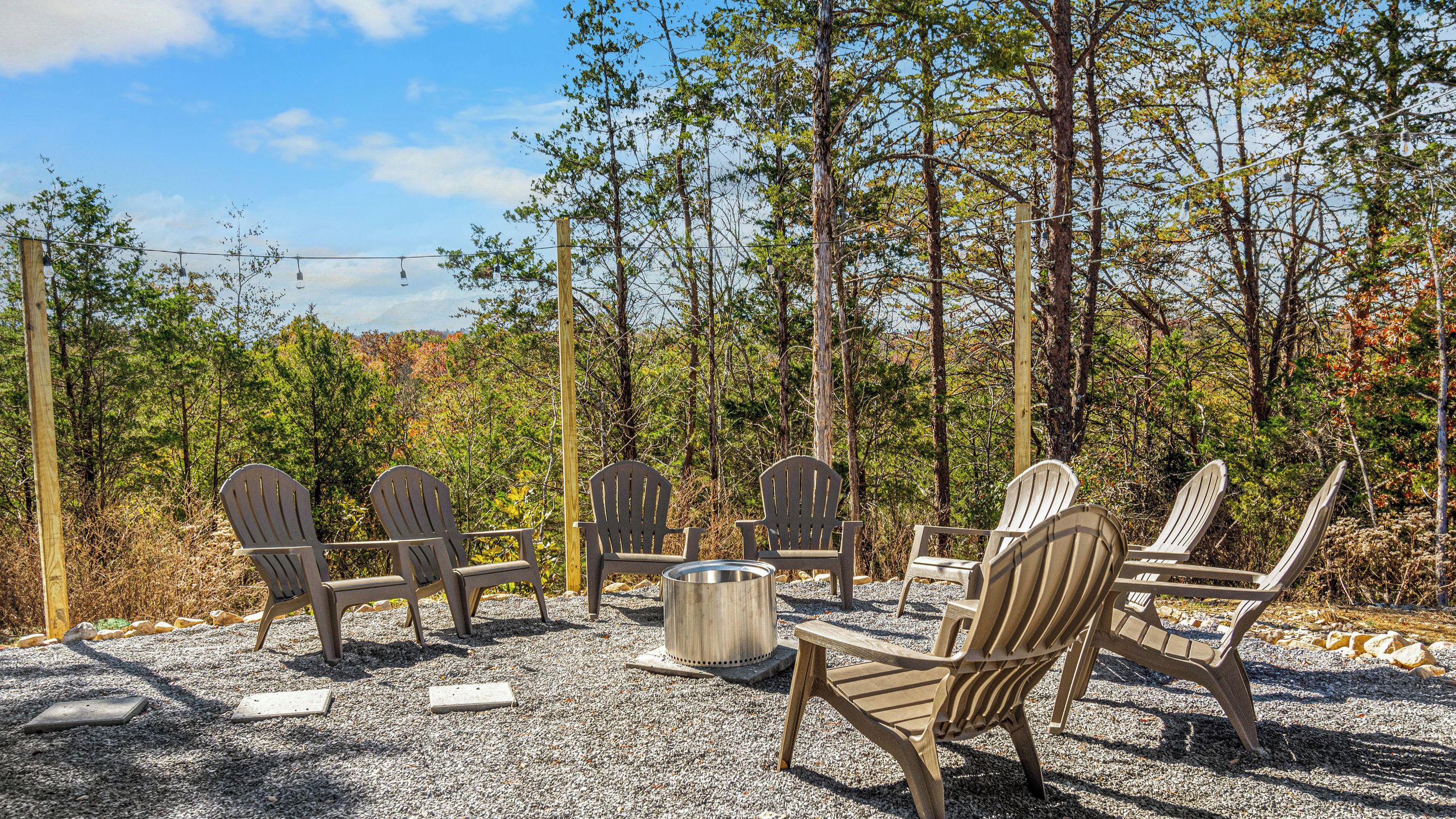A seating area with Adirondack chairs and a metal fire pit surrounded by gravel, set against a backdrop of trees and a clear sky.