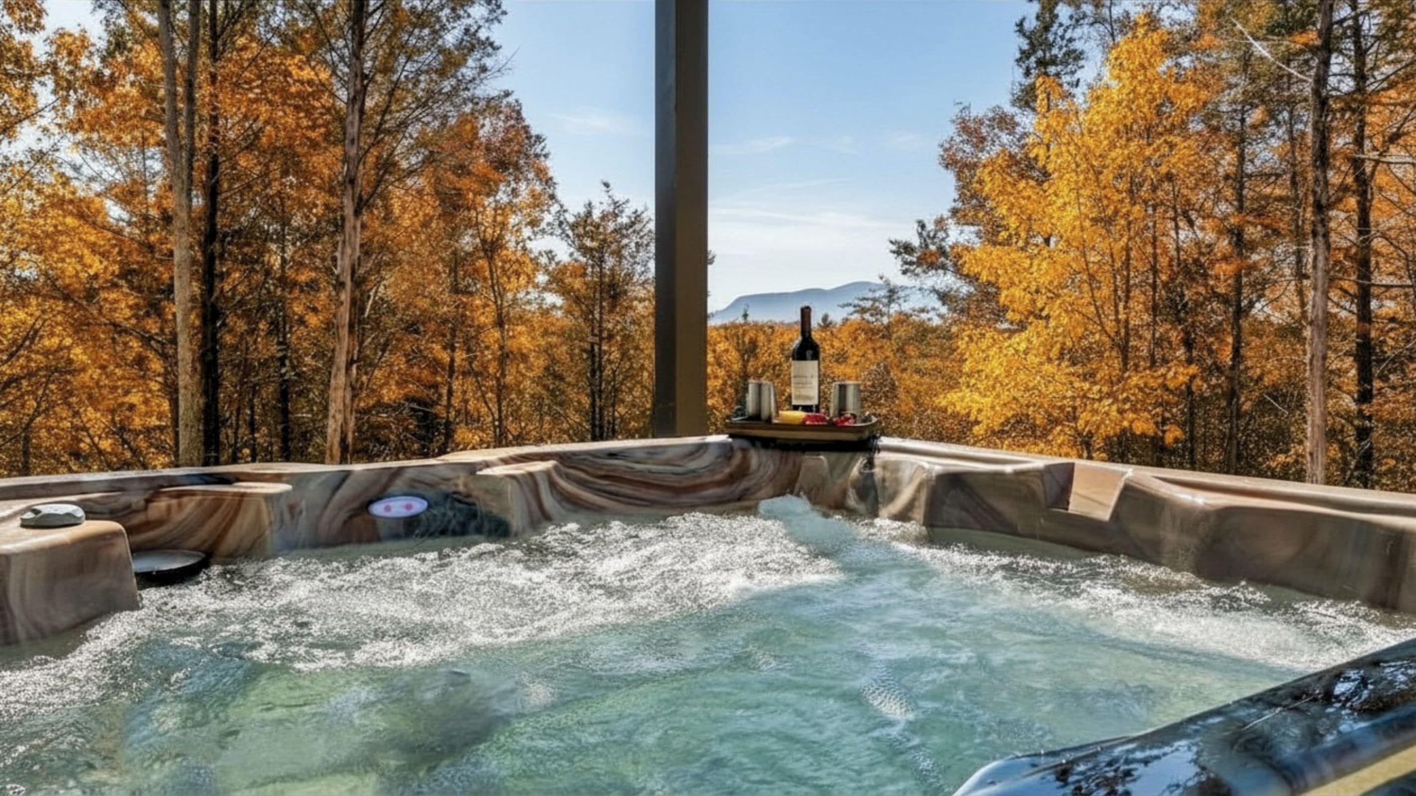 A hot tub surrounded by autumn trees with a bottle and glasses on a tray.