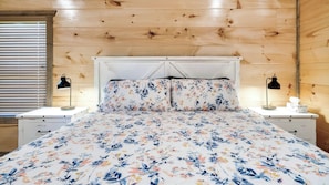 A bedroom with a wooden ceiling, a bed with floral bedding, two bedside tables with lamps, and a window with blinds. - Cabin Bliss | Hot Tub, Games & Scenic Yard Escape (Sevierville)