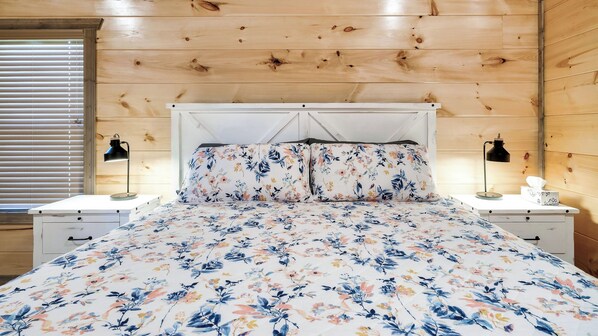 A bedroom with a wooden ceiling, a bed with floral bedding, two bedside tables with lamps, and a window with blinds.