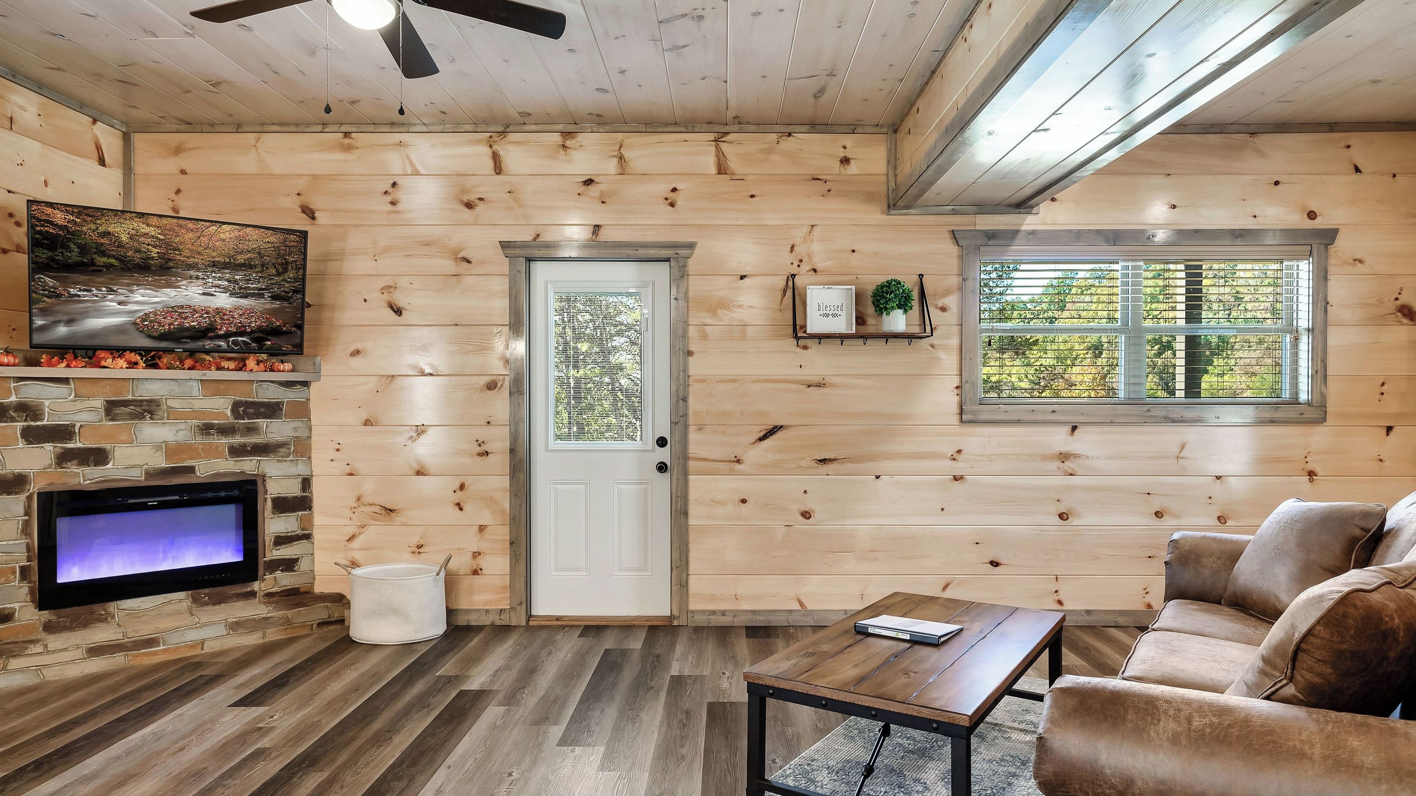 A cozy cabin interior with a stone fireplace, wooden walls, a television, a sofa, and a window with blinds.
