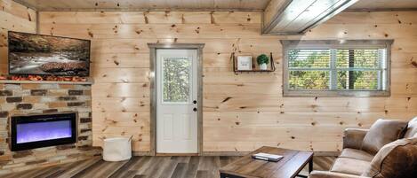 A cozy cabin interior with a stone fireplace, wooden walls, a television, a sofa, and a window with blinds.