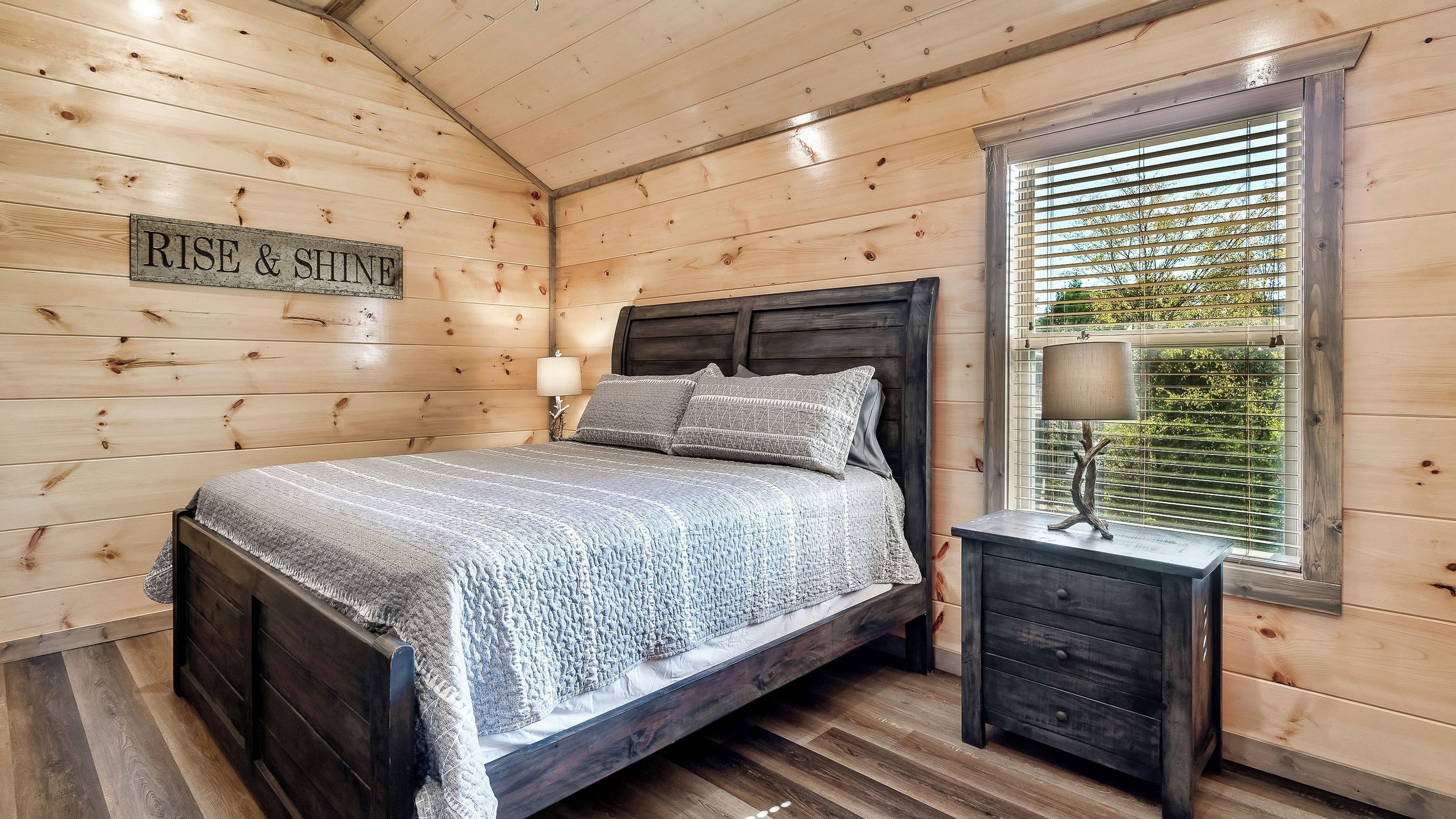 A wooden cabin bedroom with a bed, a nightstand, a lamp, and a window with blinds.