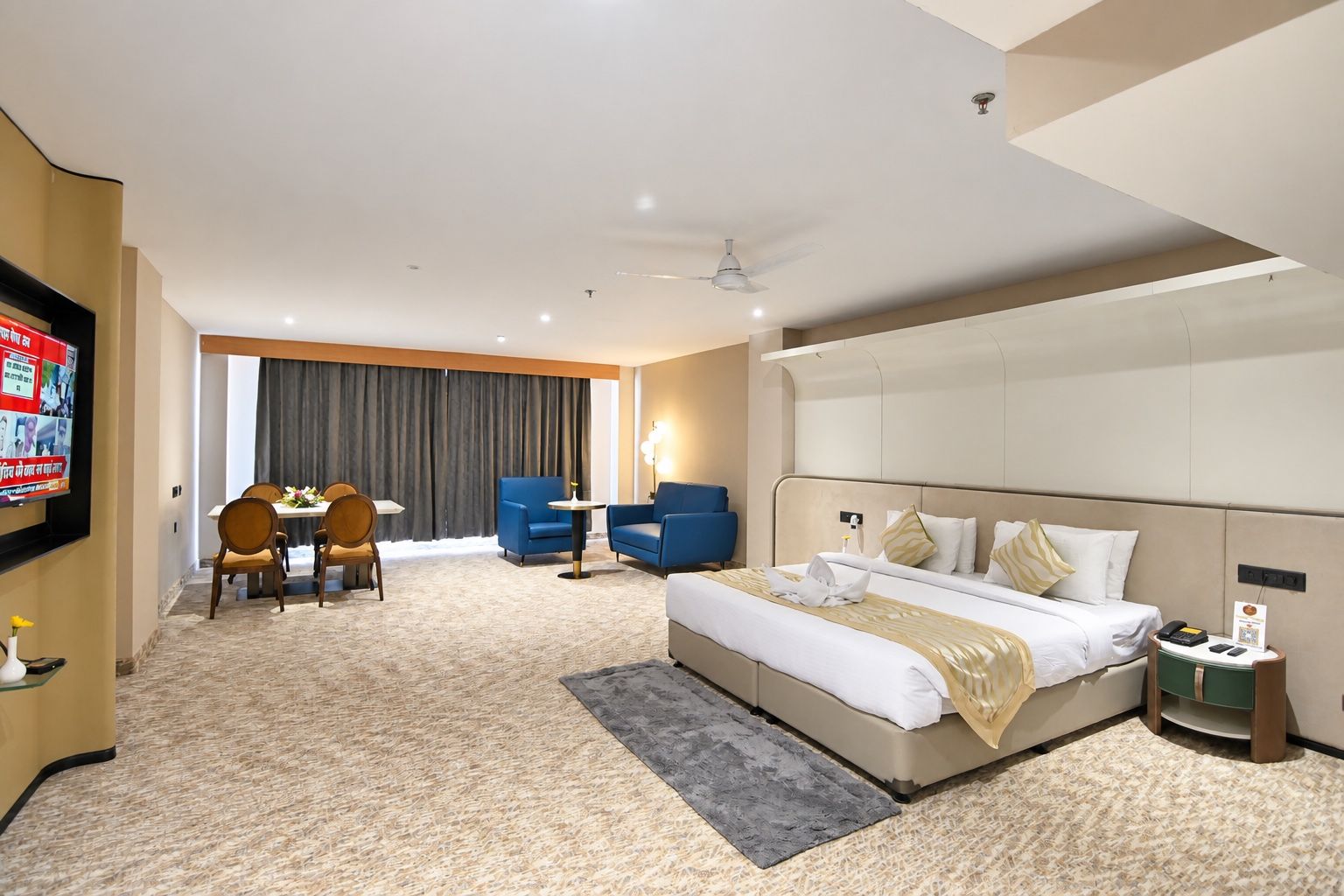 A hotel room with a bed, a desk, two chairs, a TV, and a ceiling fan.