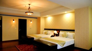 A hotel room with two beds, a desk, a chair, a red rug, and a ceiling fan. - Trinity Grand (Raigarh)
