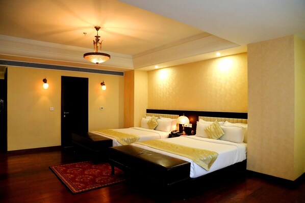 A hotel room with two beds, a desk, a chair, a red rug, and a ceiling fan. - Trinity Grand  (Raigarh)