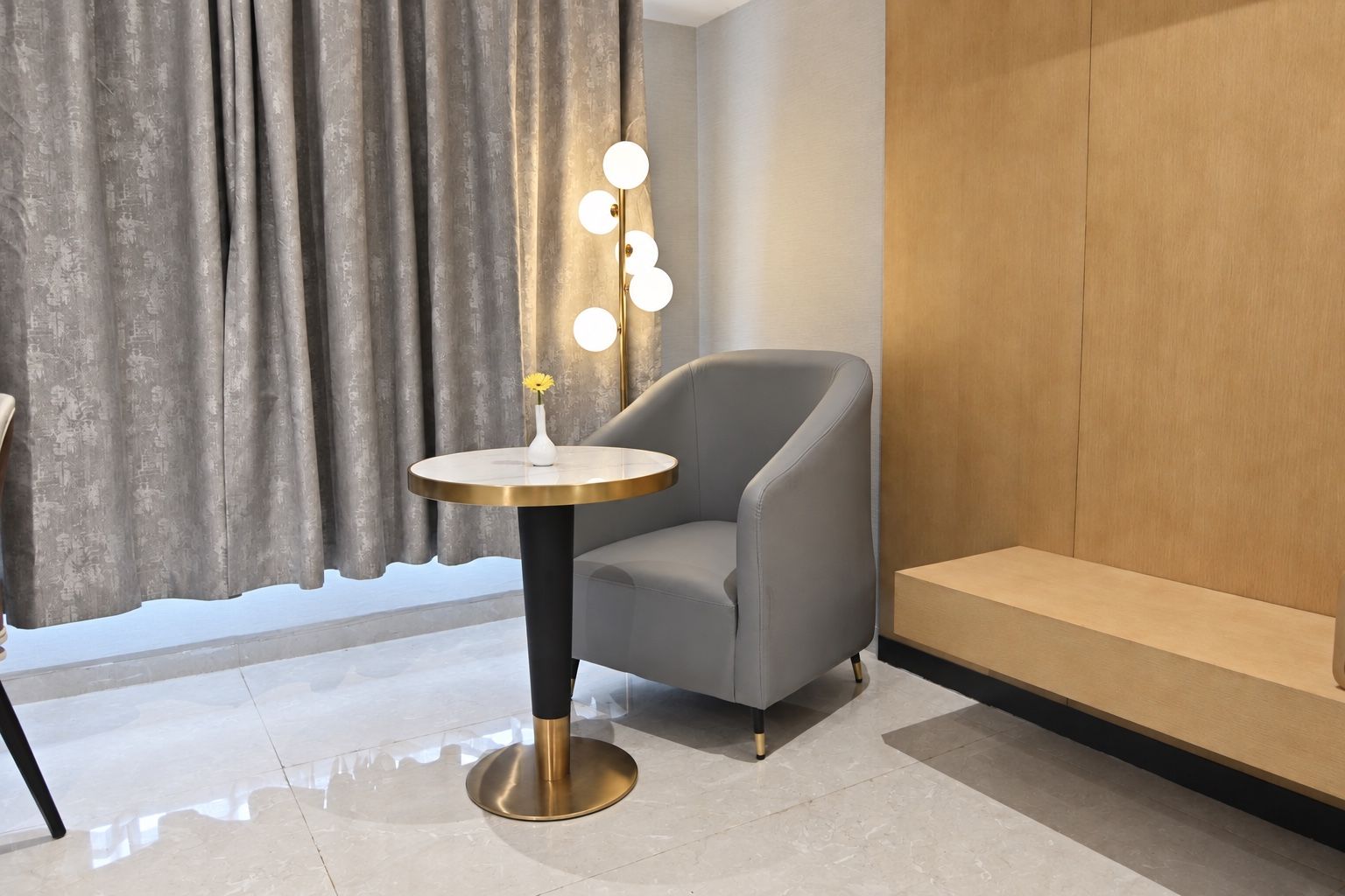 Ein modernes Zimmer mit einem grauen Sessel, einem runden Tisch mit einer Vase und Blumen sowie einer wandmontierten Leuchte.