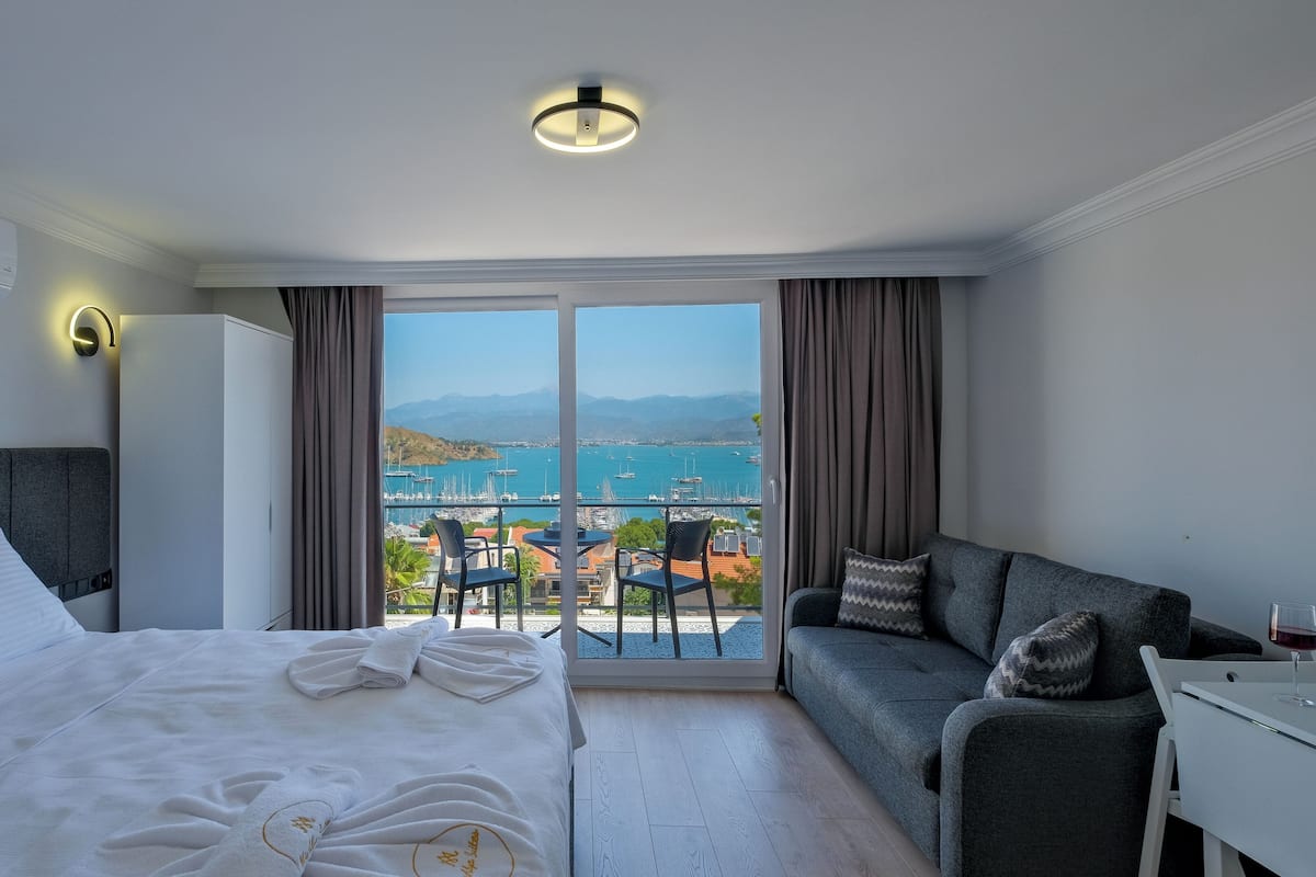 Habitación de hotel con cama, sofá, una mesa con un vaso y vistas a un puerto deportivo y montañas.