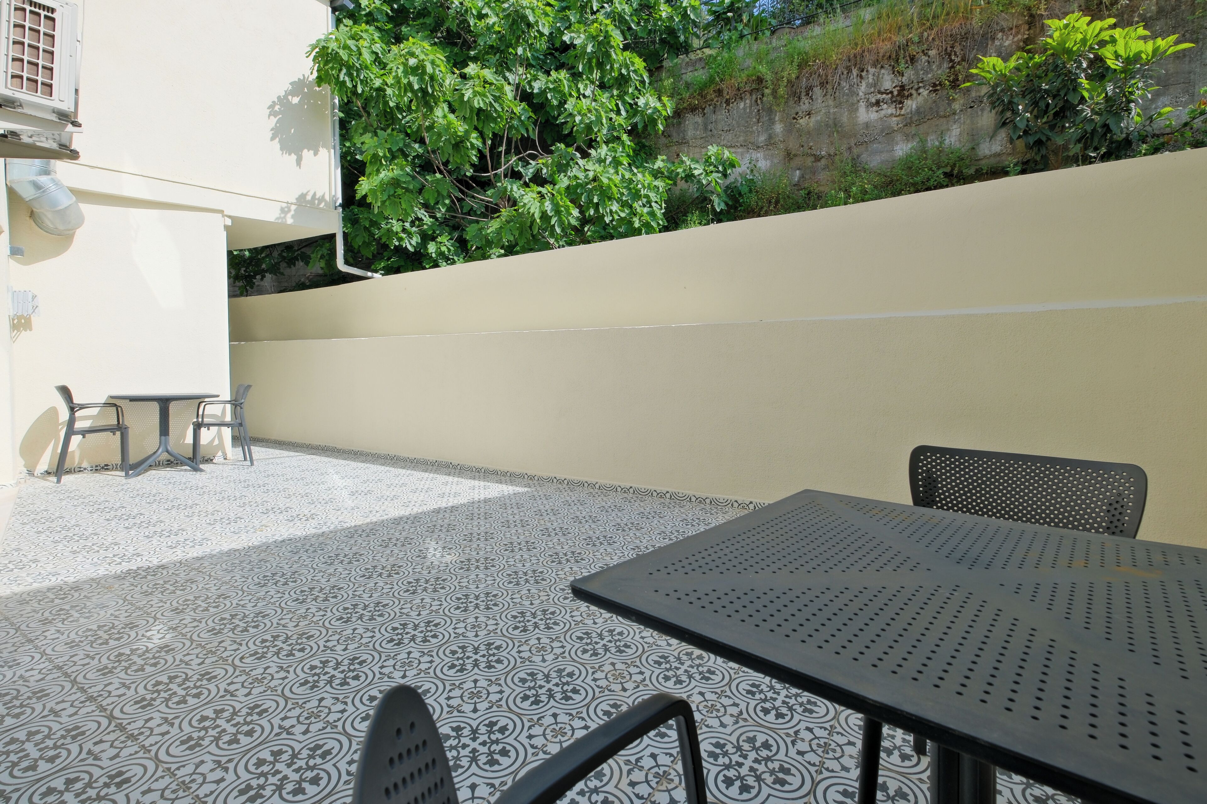 An outdoor patio with patterned flooring, a table, chairs, and a wall with greenery.