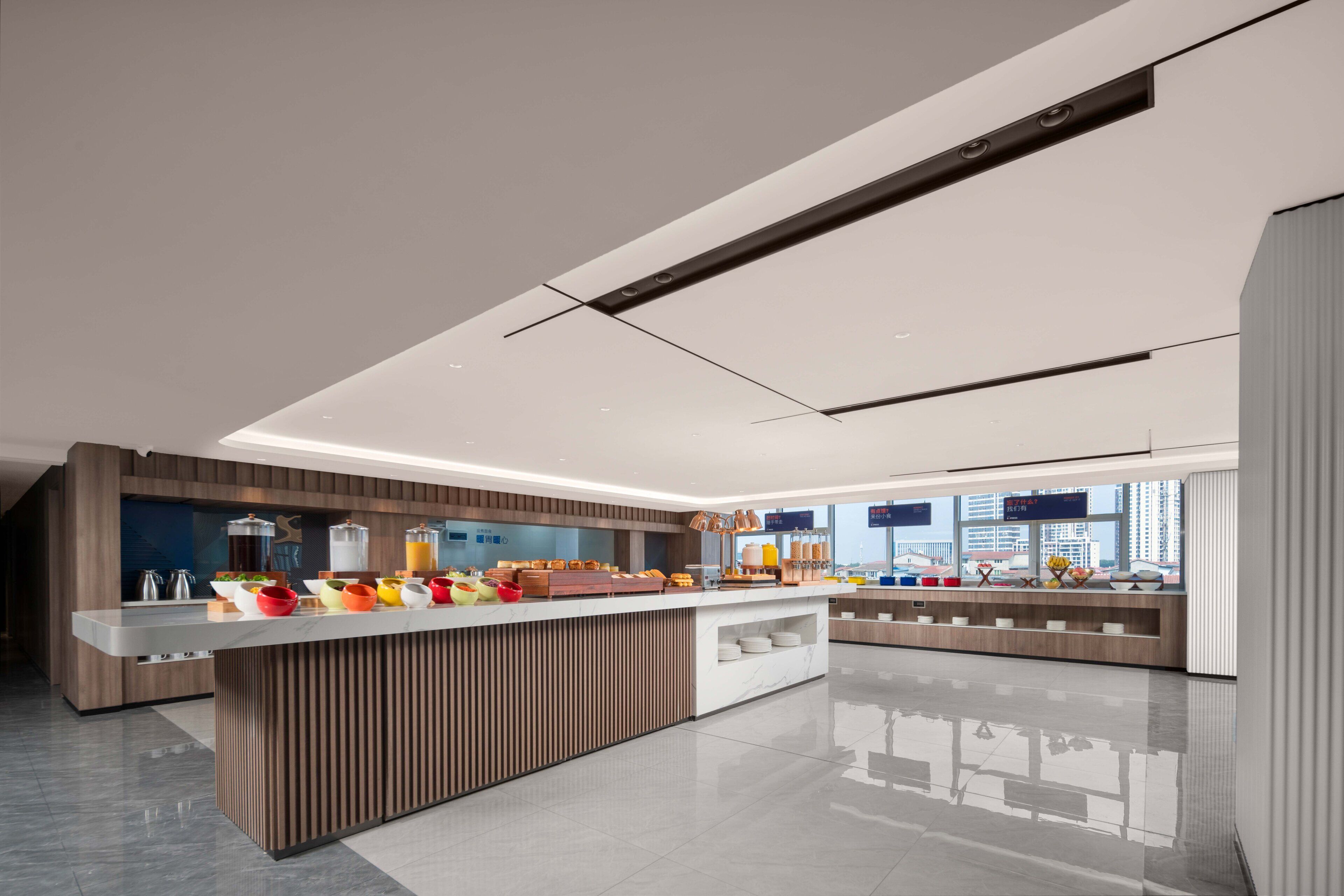 A modern hotel lobby with a central breakfast bar, wooden paneling, and a view of the cityscape through large windows.