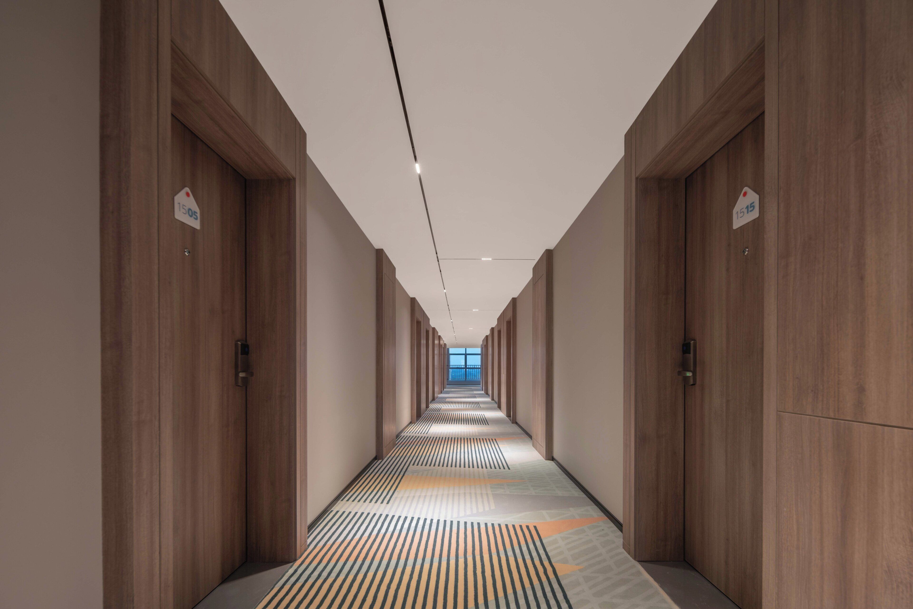 A long hallway with wooden doors, each marked with a number. The floor has a patterned carpet.