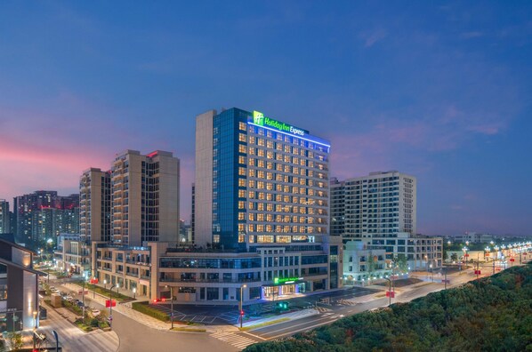 A Holiday Inn Express hotel with a modern design, surrounded by other buildings and a well-lit street at dusk. - Holiday Inn Express Mianyang Sci-Tech City by IHG (Mianyang)