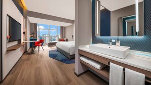 Een moderne hotelkamer met een groot bed, een bureau met stoel, een grote spiegel en een badkamer met een wit wastafelmeubel en handdoekhouder.