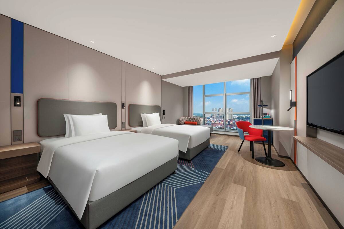 Ein Hotelzimmer mit zwei Betten, einem Schreibtisch, einem Sessel und einem großen Fenster mit Blick auf die Stadt.