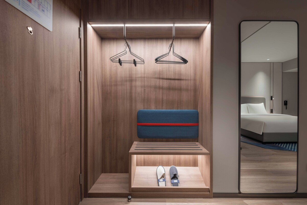 A modern hotel room with a wooden wardrobe, a built-in shoe rack, and a mirror reflecting a bed and a bathroom.