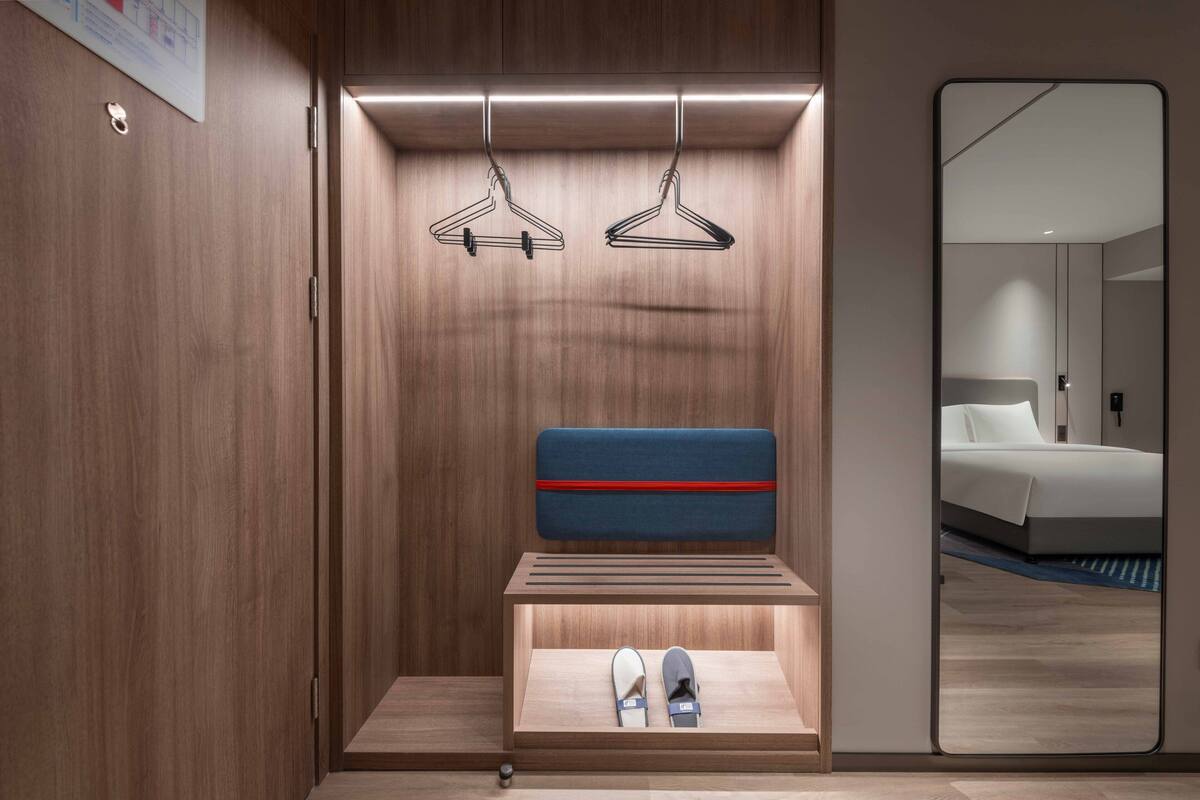 Ein modernes Hotelzimmer mit einem Holzschrank, einer eingebauten Schuhablage und einem Spiegel, der ein Bett und ein Badezimmer reflektiert.
