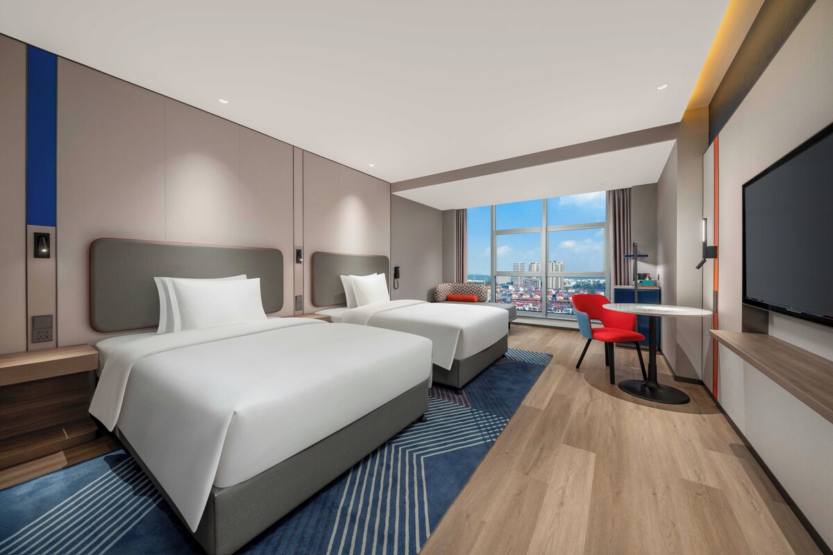 A hotel room with two beds, a desk, a chair, and a large window offering a city view.