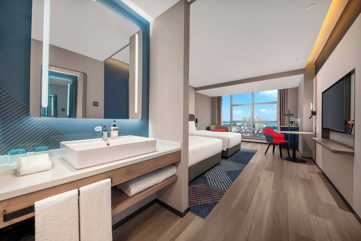 A modern hotel room with a large mirror, a white sink, a bed, a desk with a chair, and a TV.