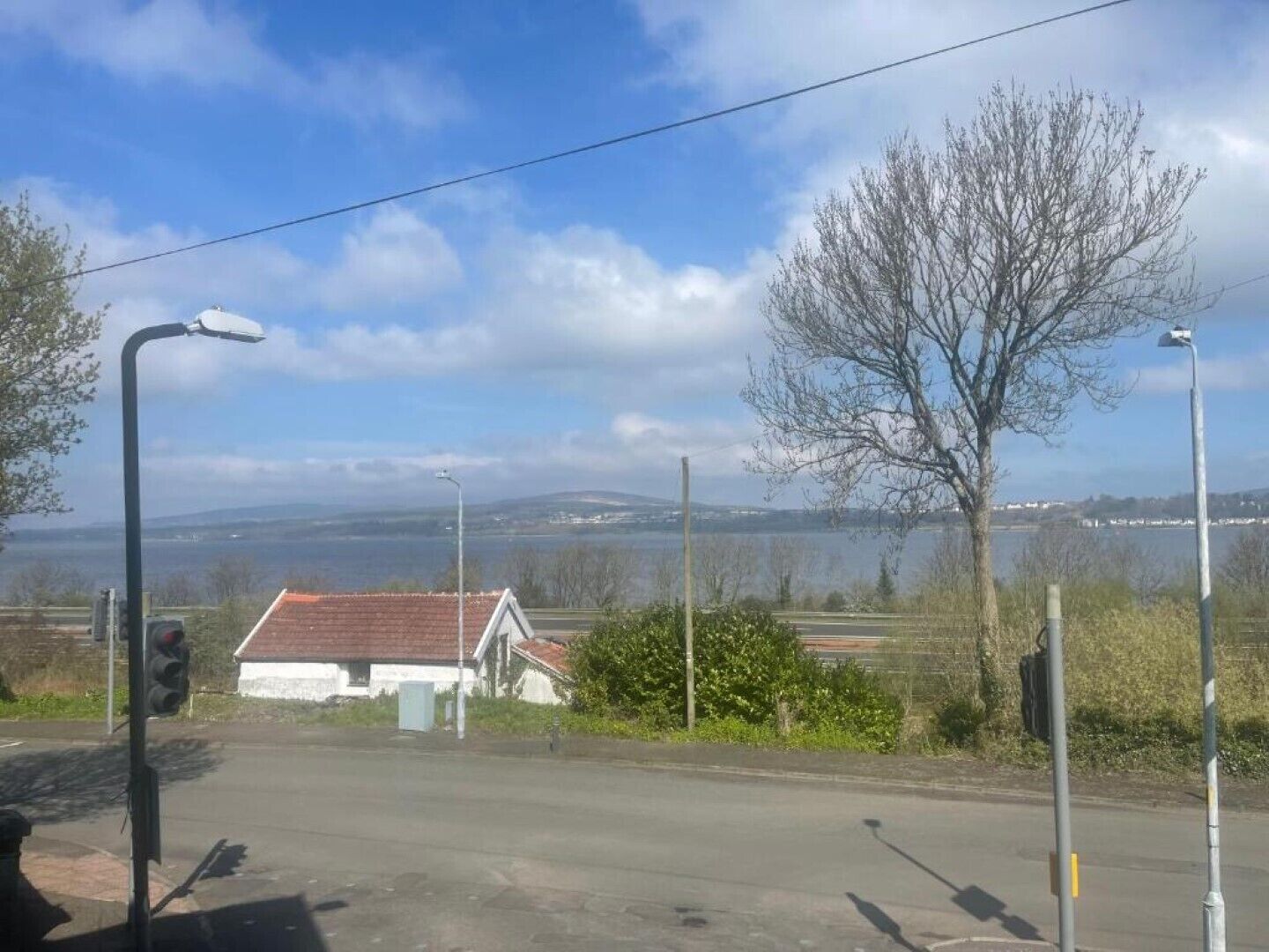 A rural road with a white building with a red roof, surrounded by trees and a view of a body of water and hills in the distance.