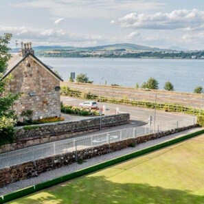 A stone building with a fenced area and a view of a body of water and distant hills. - Clydeview Apartments (Langbank)