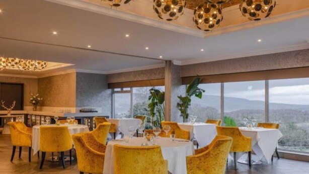 Un comedor con mesas redondas y sillas amarillas, ventanas amplias con vistas, y una lámpara de araña moderna.