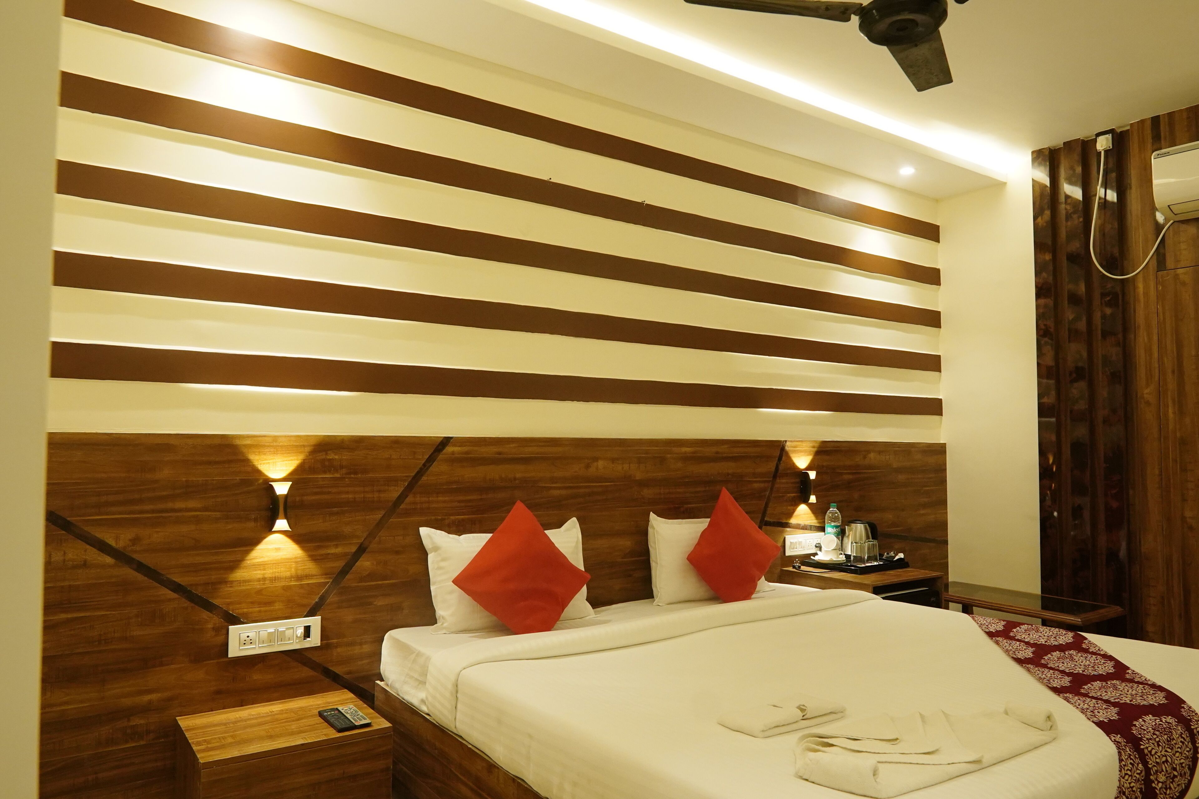 Une chambre d’hôtel avec un grand lit, une tête de lit en bois, deux tables de chevet, une petite table, un radiateur fixé au mur et un ventilateur de plafond.