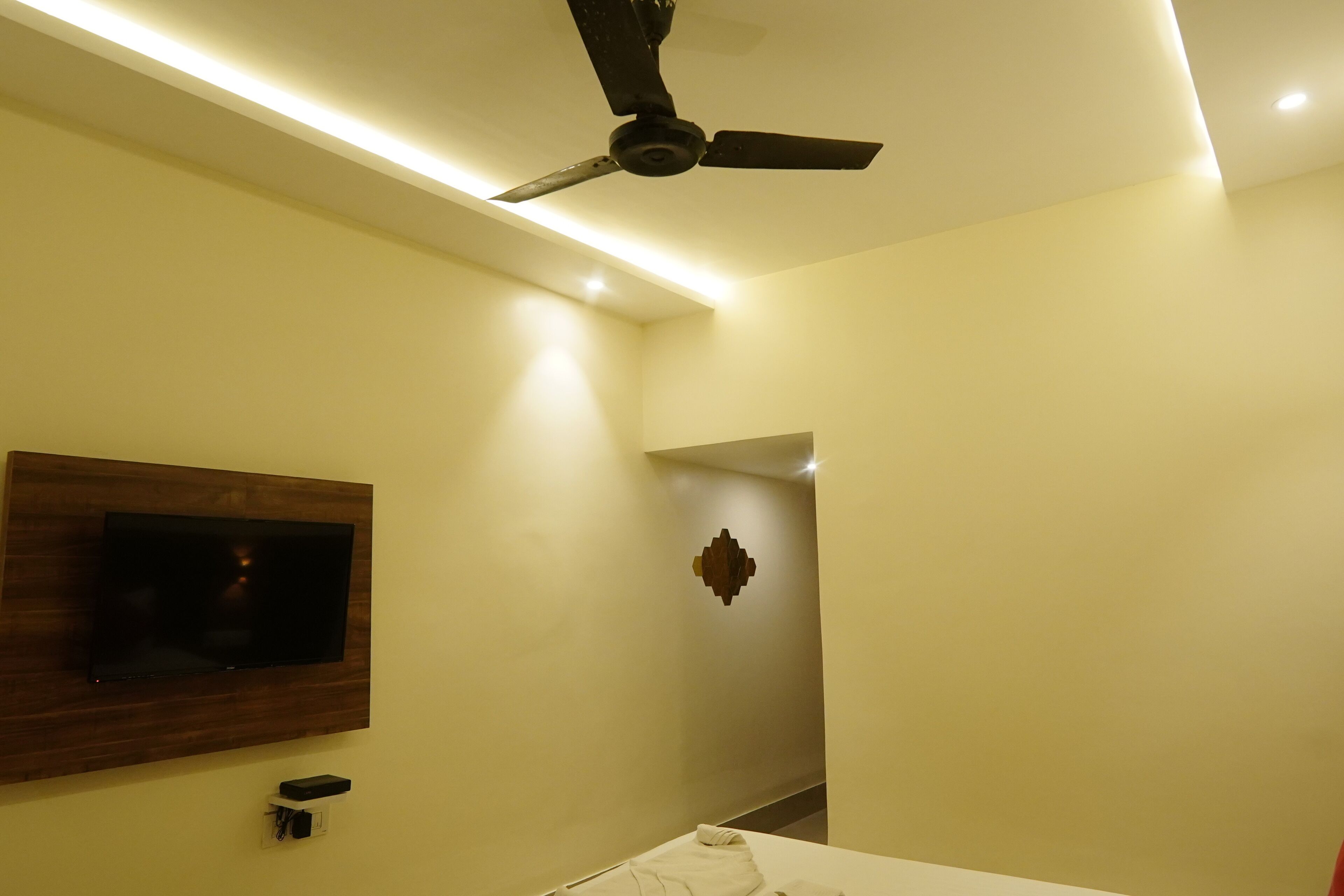 Une chambre d’hôtel équipée d’une télévision fixée au mur, d’un ventilateur de plafond et d’une petite table de chevet.