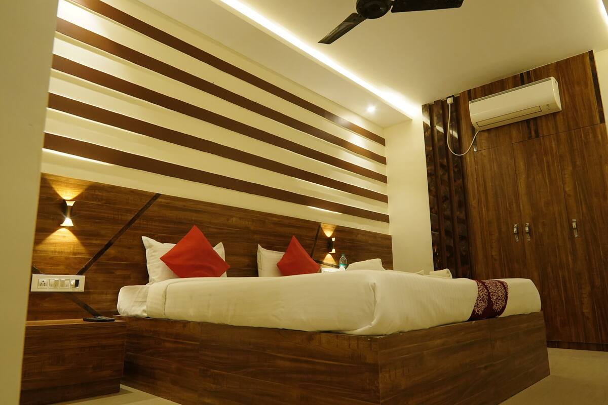 Un dormitorio moderno con cabecera de madera, cama con ropa blanca y almohadas rojas, mesita de noche, equipo de aire acondicionado y ventilador de techo.