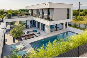 Una casa moderna de dos pisos con balcón de vidrio, piscina y mobiliario de exterior.
