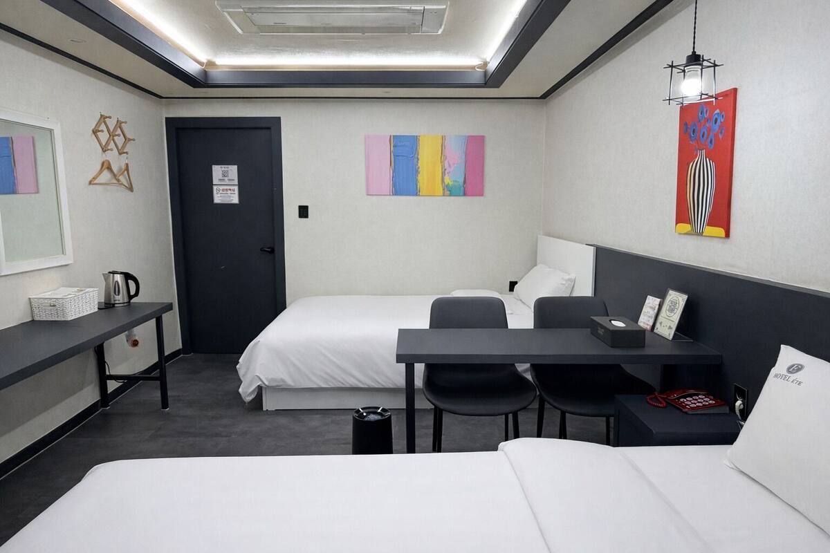 A hotel room with two beds, a desk, a chair, a small table, a kettle, and a trash bin.