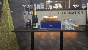 A reception area with a reception desk, a clock displaying time in Seoul, USA, and Paris, a champagne bottle, a cake, and a blue box with a logo. - Hotel Carlton Incheon Airport Juan (Incheon)