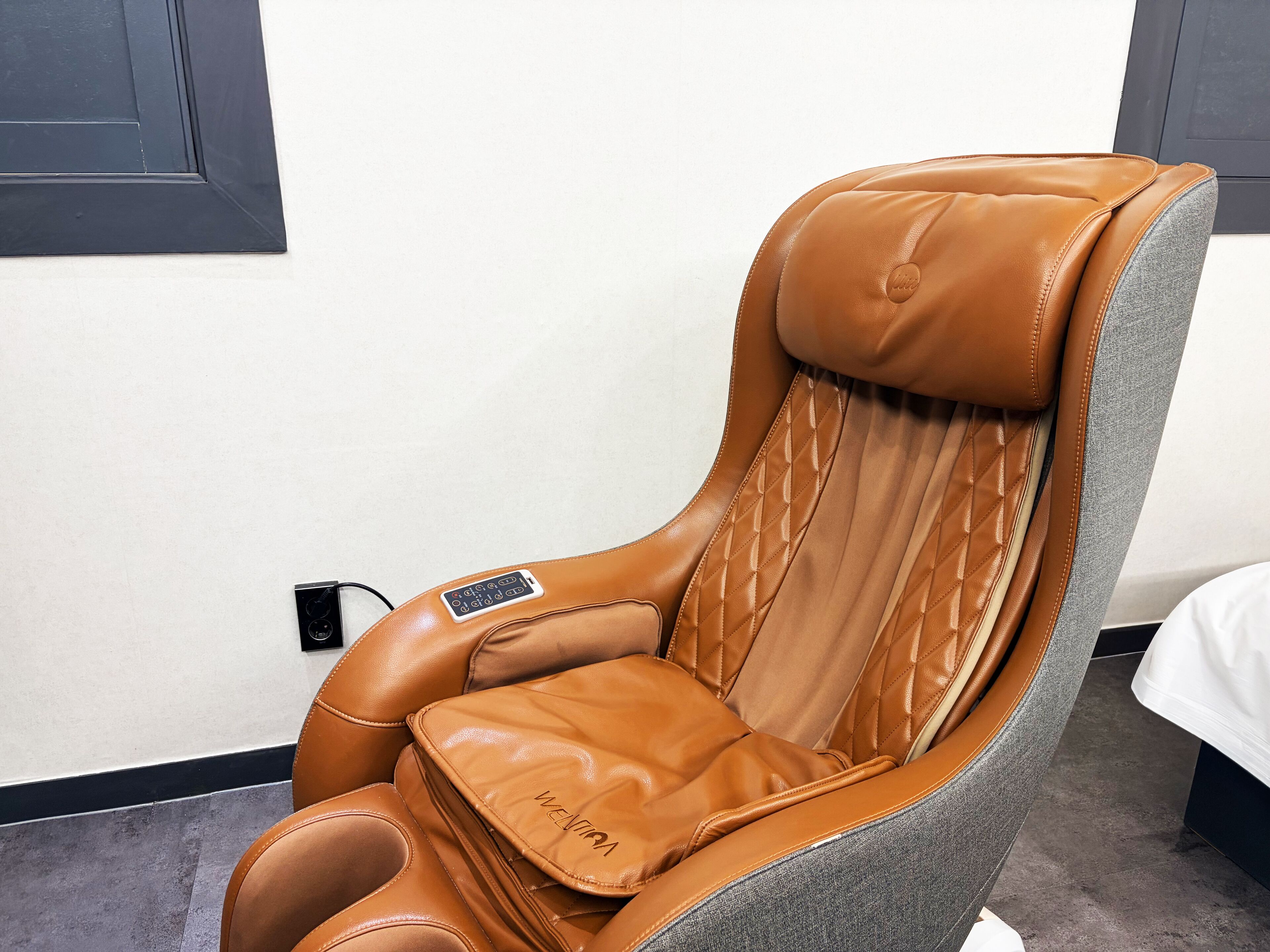A massage chair with a control panel, positioned against a white wall with a black door frame.