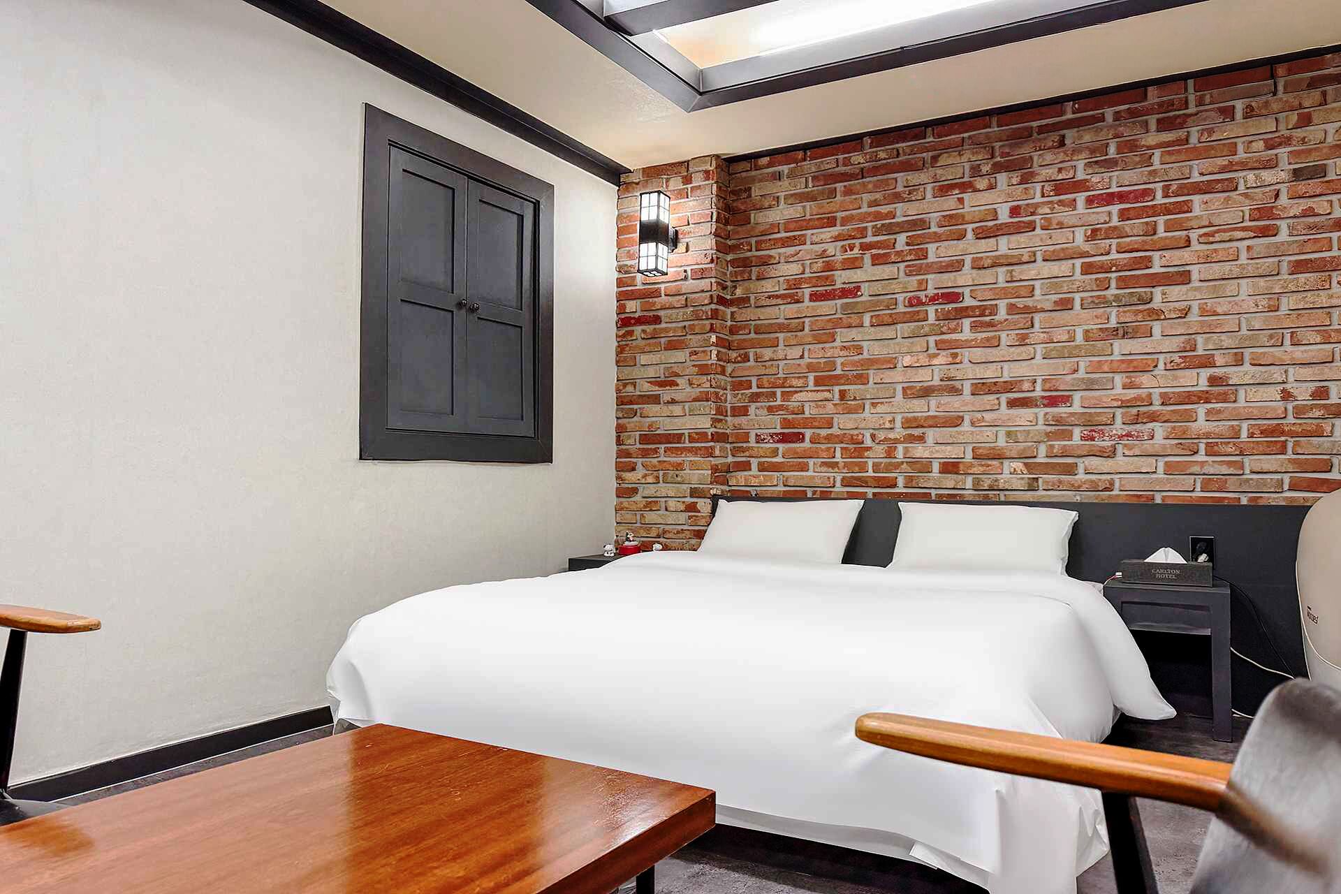 A hotel room with a brick wall, a bed with white bedding, a nightstand, and a wooden chair.