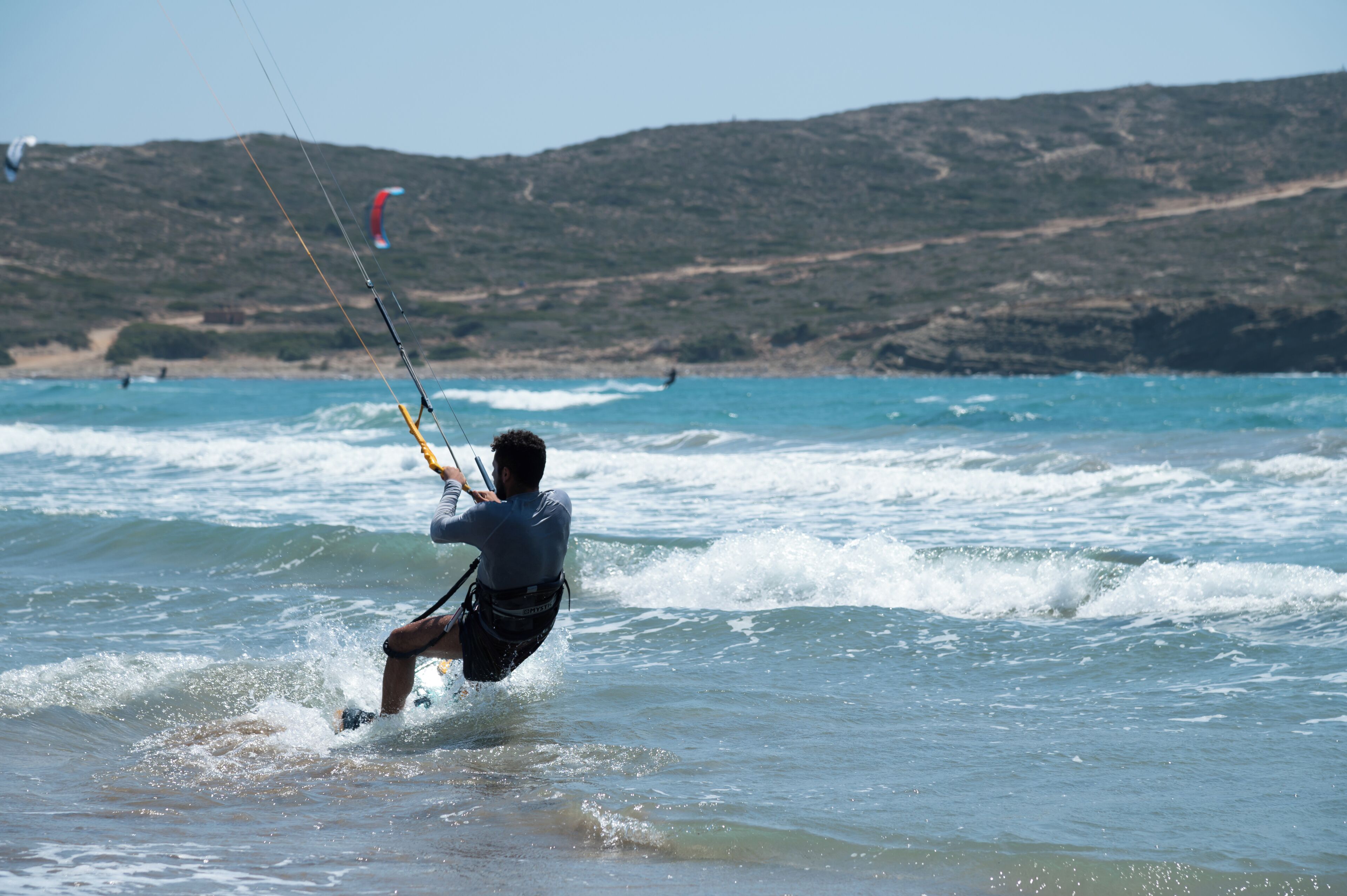 A person windsurfing on a beach with waves and a hilly landscape in the background.