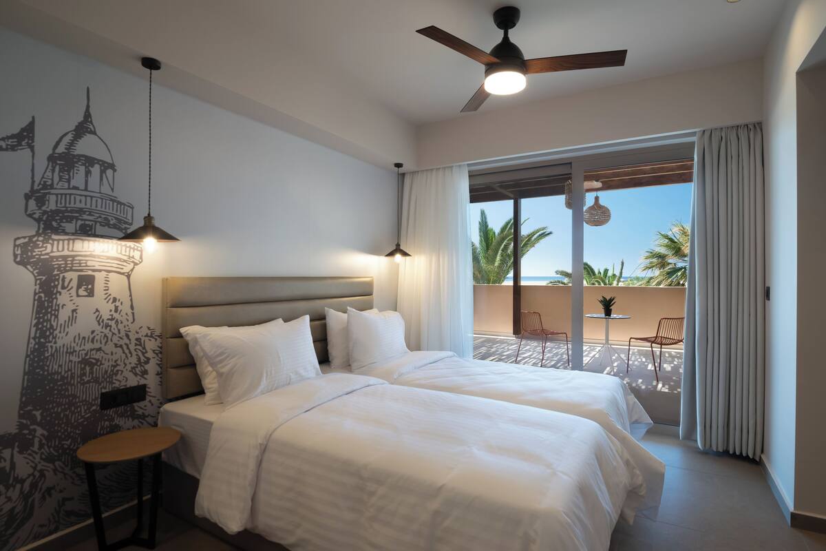 Ein Hotelzimmer mit zwei Betten, einem Deckenventilator, einem Balkon mit Blick auf Palmen und einem Wandgemälde.