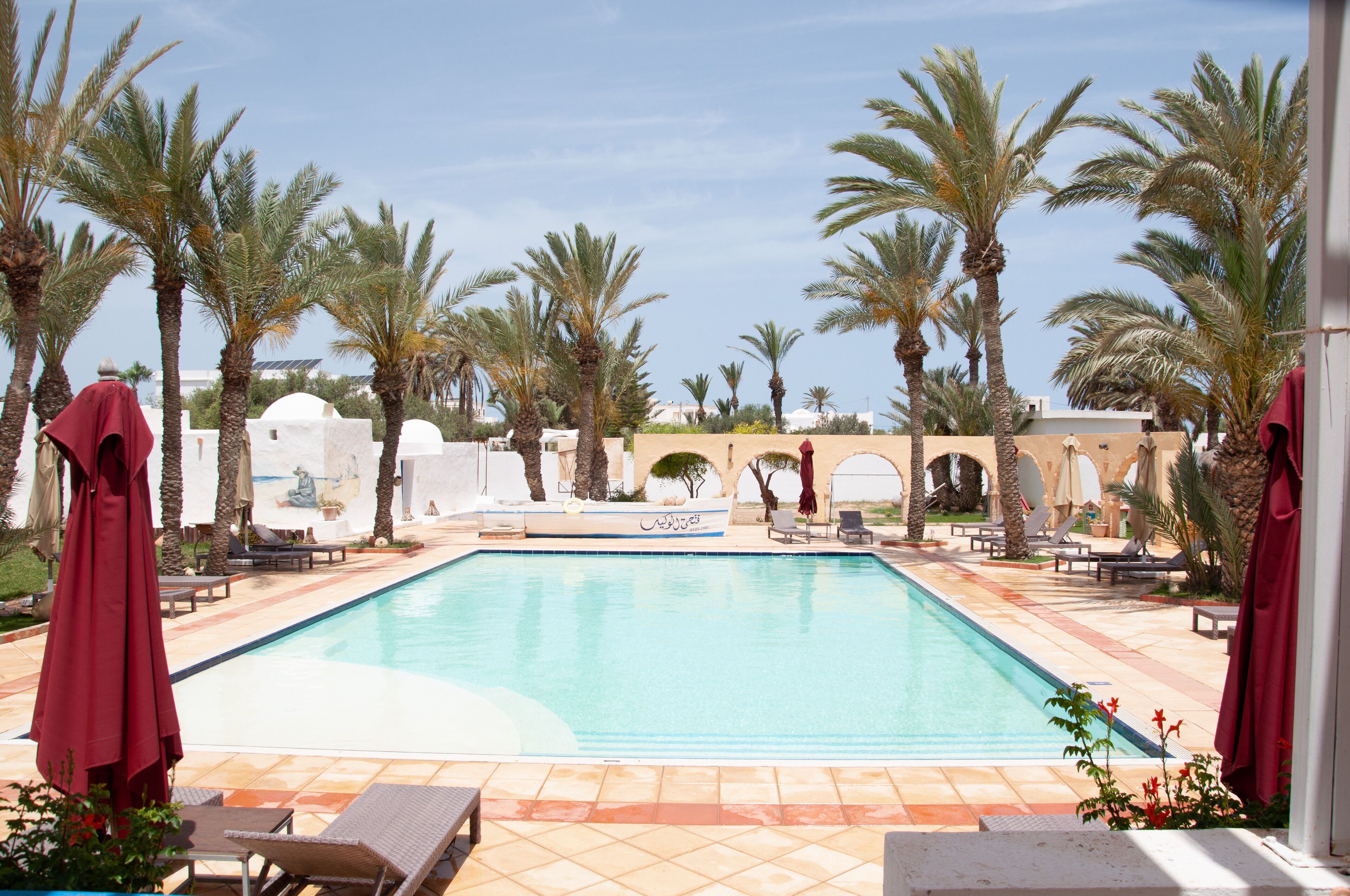 A swimming pool with lounge chairs and umbrellas, surrounded by palm trees and a white building with arches.