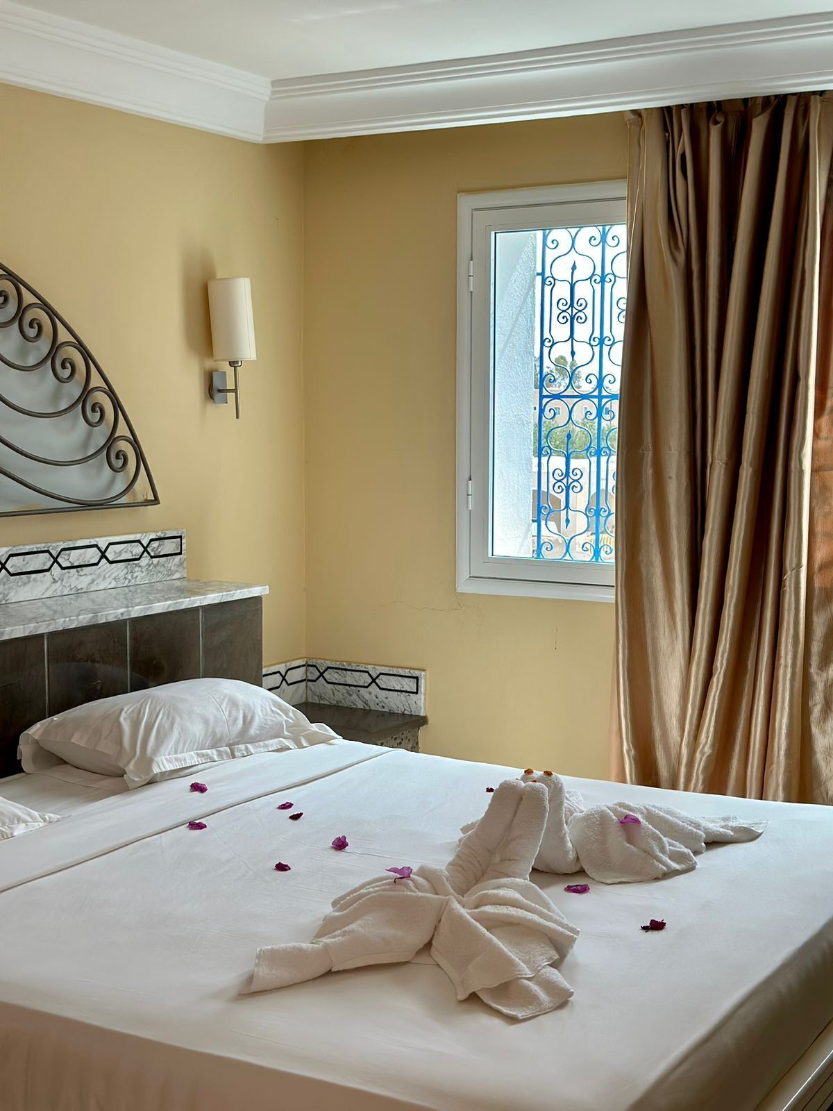 A hotel room with a bed, a nightstand, a wall-mounted light, a window with curtains, and a decorative metal railing.