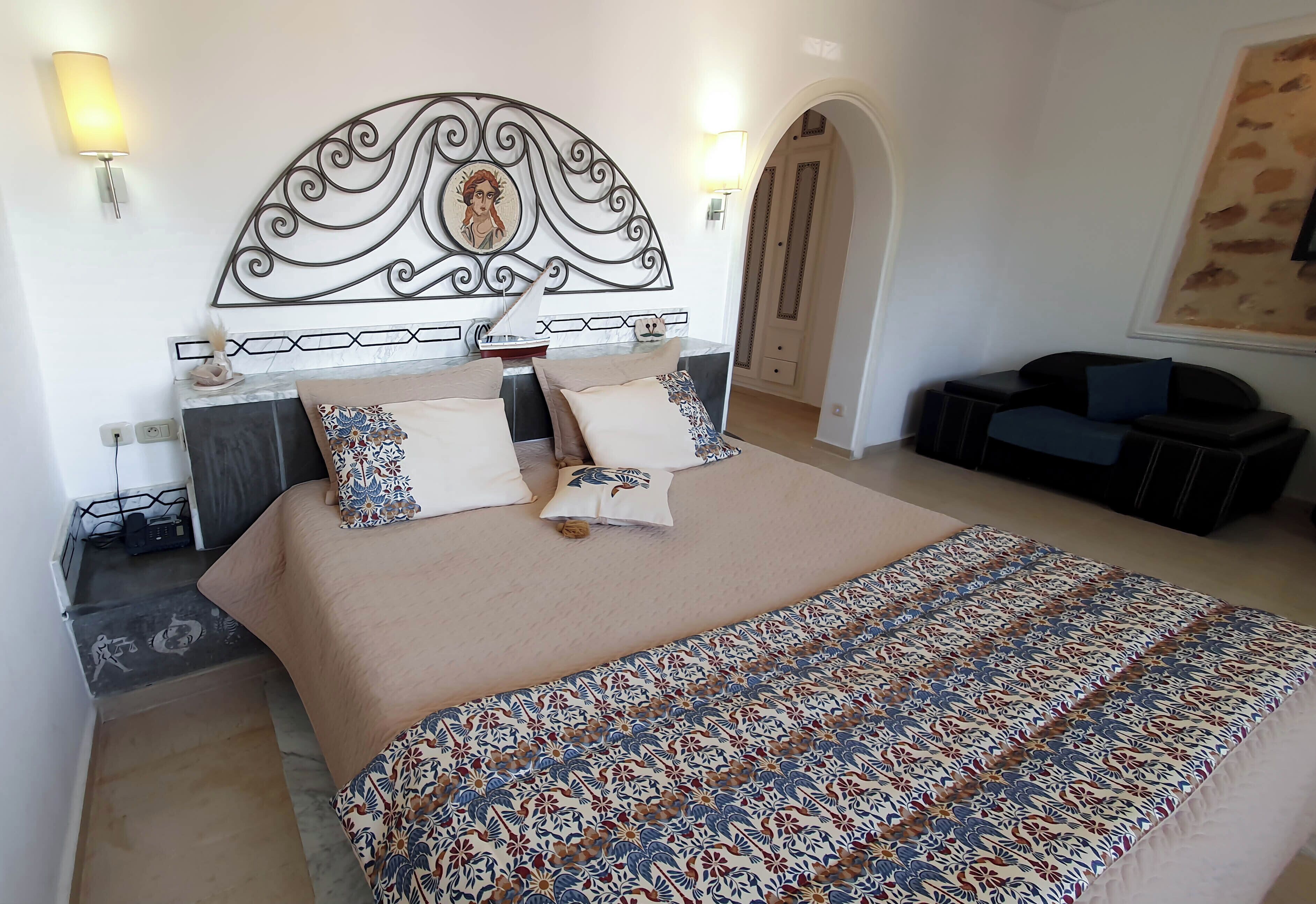 Habitación de hotel con una cama grande, un cabecero decorativo, dos lámparas fijadas a la pared, una pequeña zona de estar y una puerta que da a otra habitación.