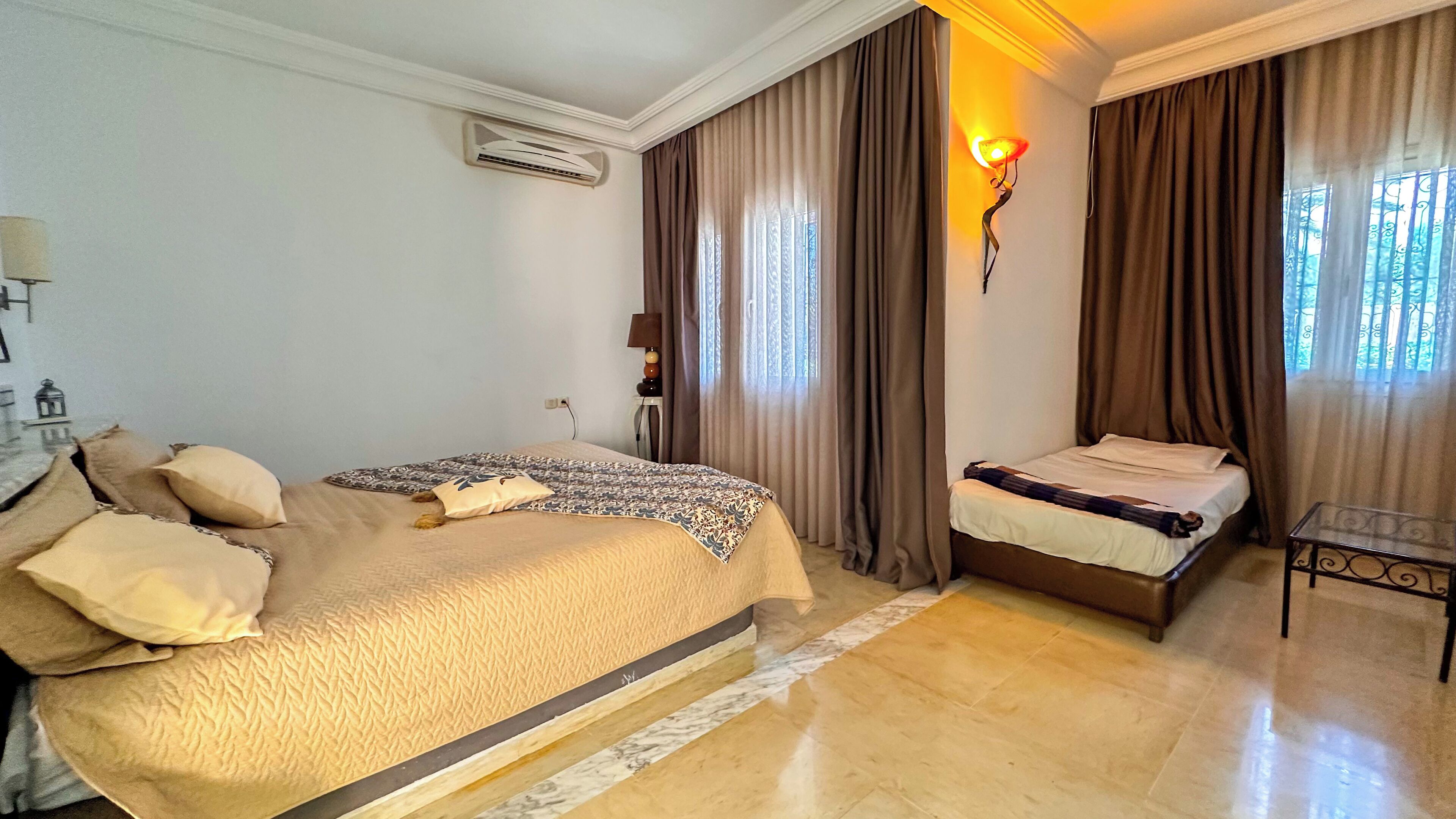A hotel room with two beds, a marble floor, a wall-mounted air conditioner, and a window with curtains.