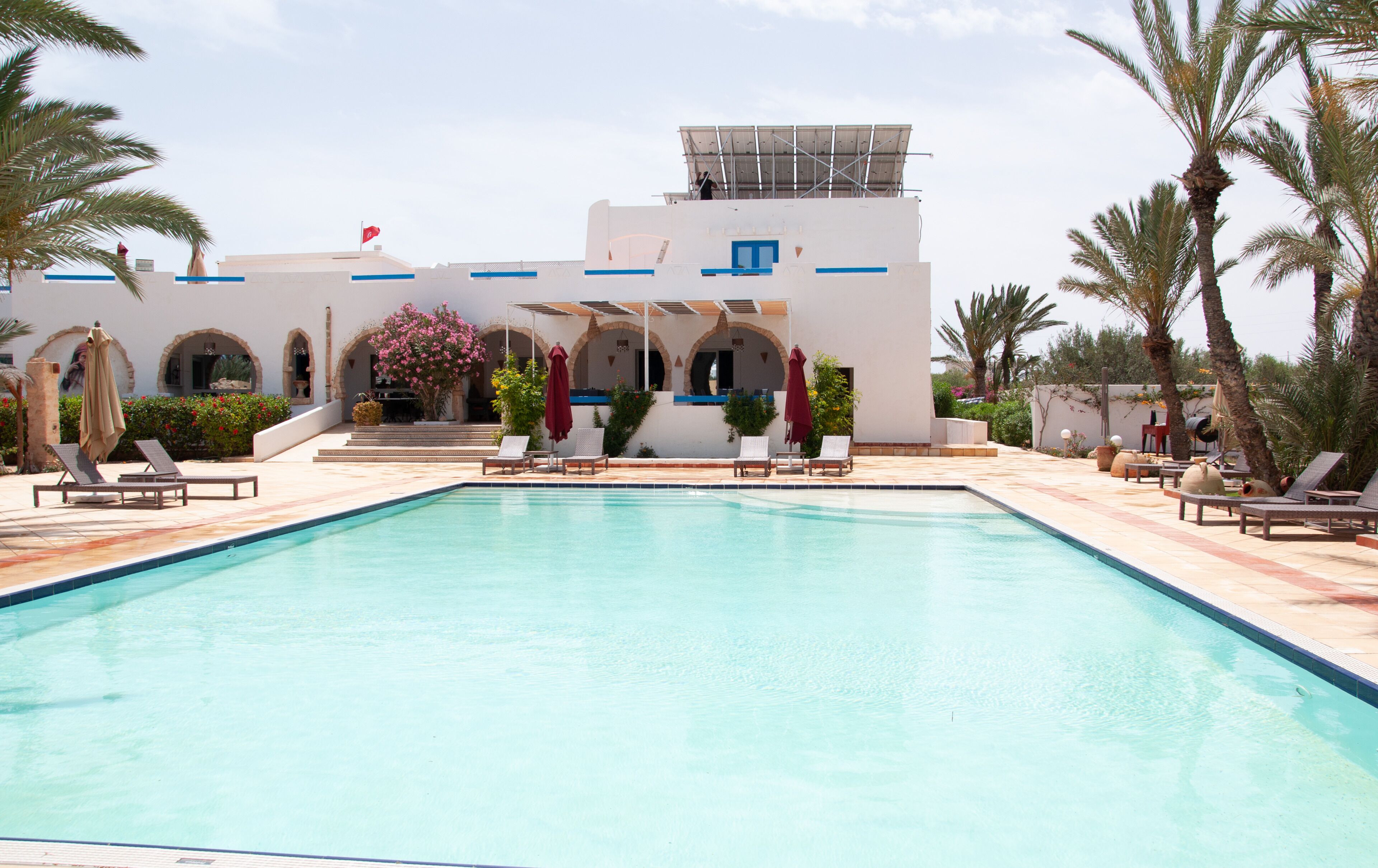 Una piscina con sillones de descanso y sombrillas, rodeada de palmeras y un edificio con arcos y una bandera.