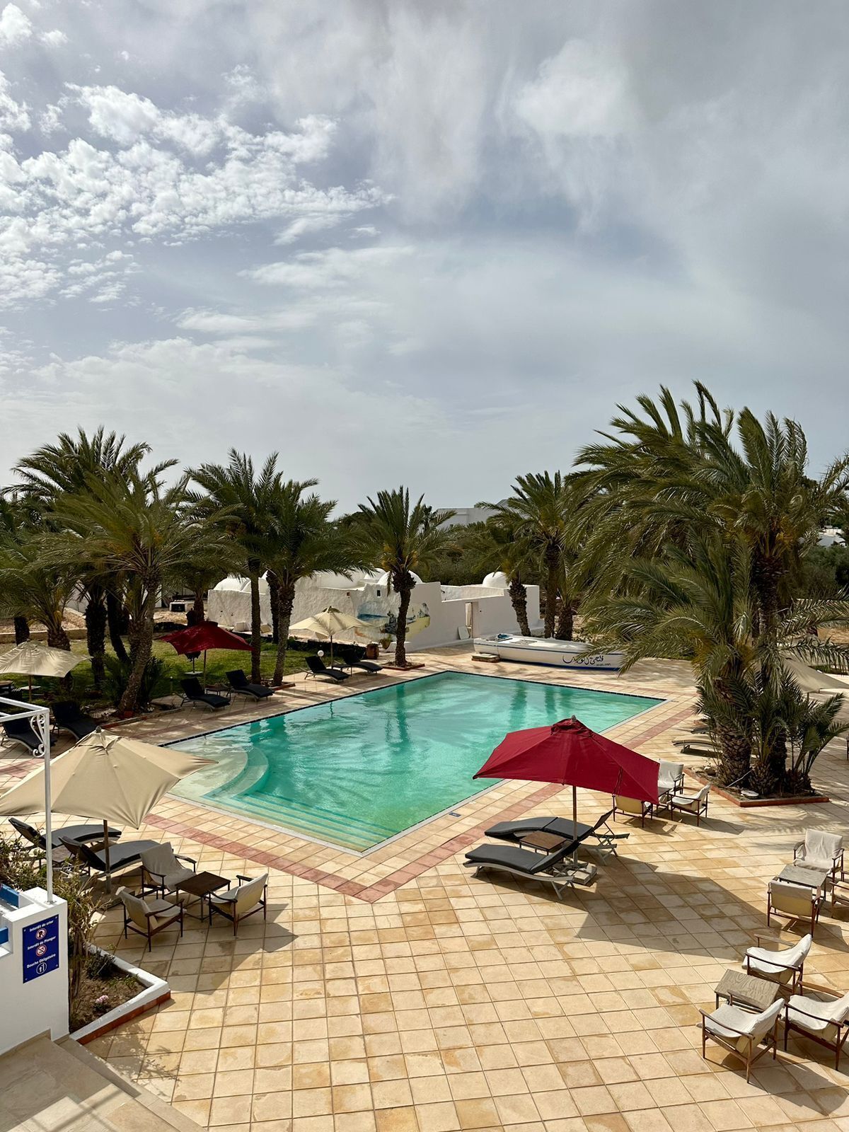 A swimming pool with lounge chairs and umbrellas, surrounded by palm trees and a tiled deck.