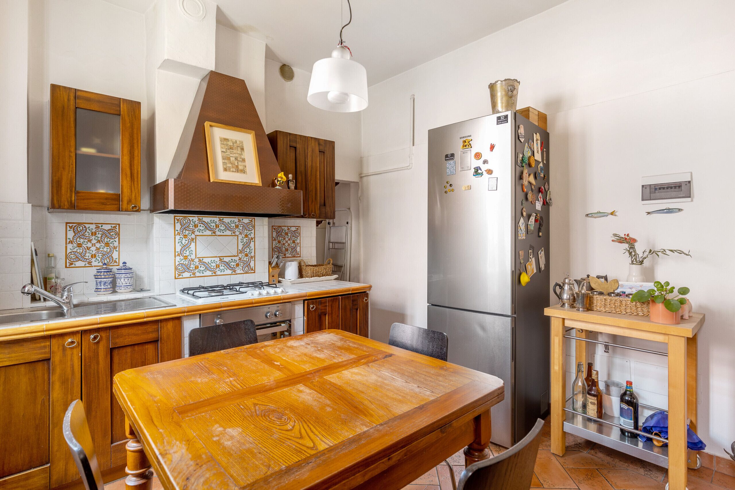 A kitchen with wooden cabinets, a stainless steel refrigerator, a wooden dining table, and a small shelf with bottles.