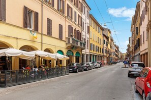 Una calle con coches estacionados, mesas al aire libre en un restaurante y edificios con balcones.
