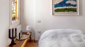 Ein Schlafzimmer mit einem Bett, einem Fenster mit Blick auf Pflanzen, einem Bild an der Wand und einem Nachttisch mit einer Lampe.