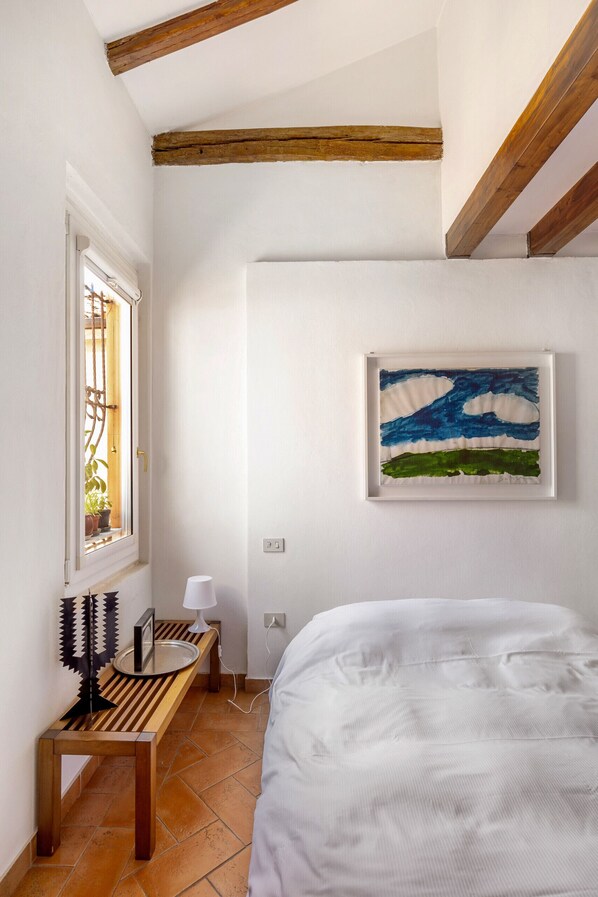 Un dormitorio con cama, ventana con vista a plantas, un cuadro en la pared y una mesita de noche con lámpara.