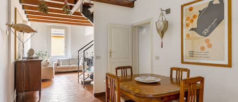 Un comedor con una mesa y sillas de madera, una escalera con barandilla de hierro forjado, y vistas a una sala de estar con sofá y ventana.