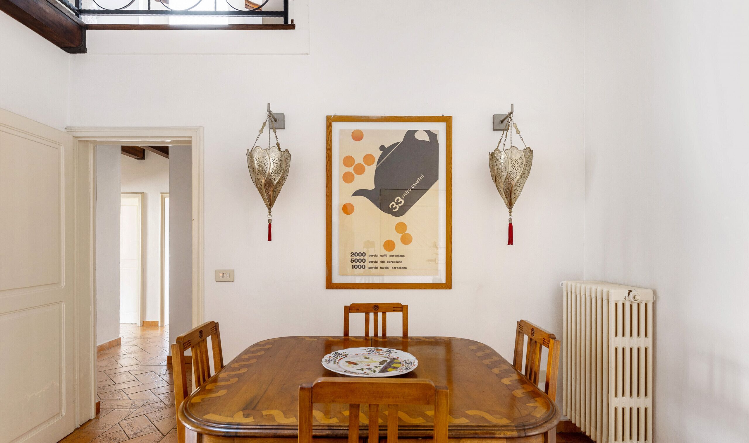 Een eetgedeelte met een houten tafel, vier stoelen, een lijstwerkkunstwerk, twee wandlampen en een radiator.