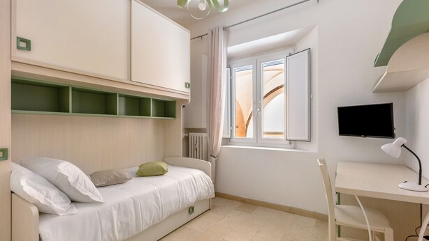 Een slaapkamer met een bed, een bureau, een stoel, een televisie en een raam met gordijnen.